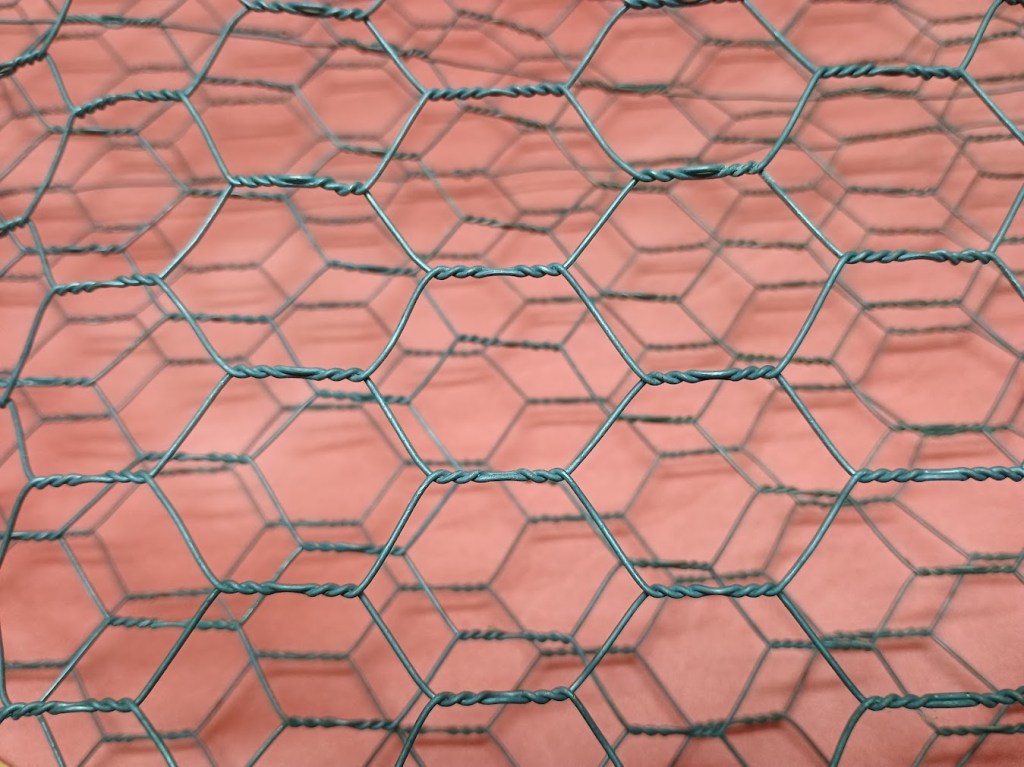 Close-up view of green chicken wire fencing with hexagonal mesh pattern against a pink background.