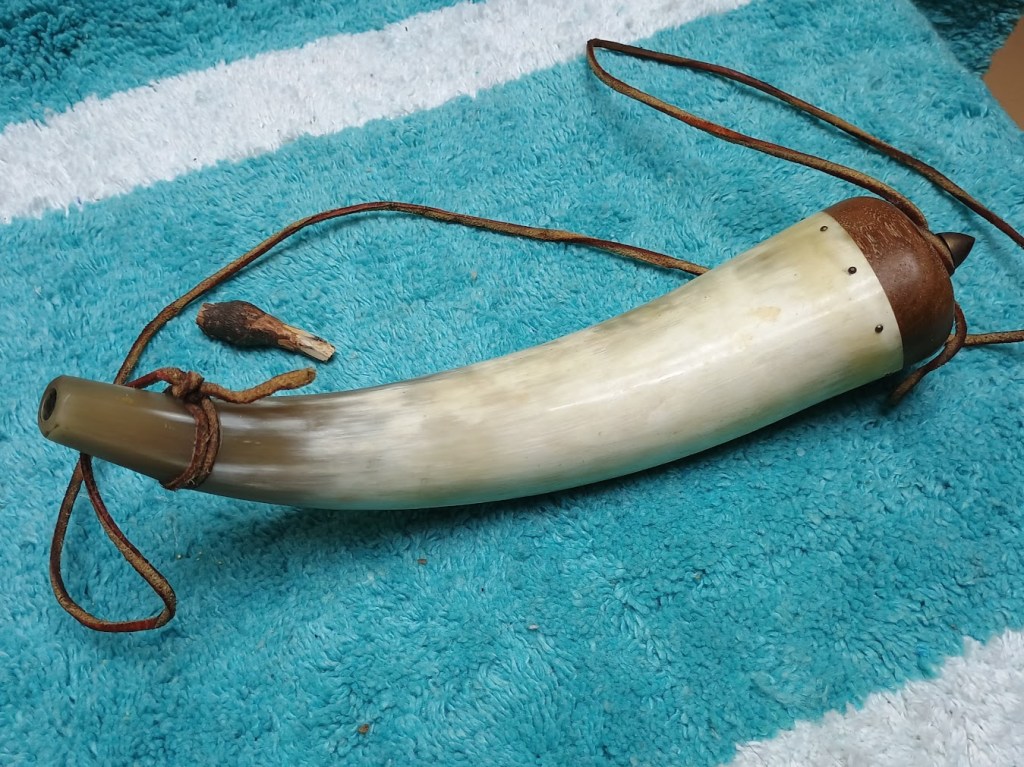 A hand-carved powder horn made from a hollowed-out animal horn, featuring a rounded top secured with a wooden stopper, and attached leather straps, placed on a textured blue surface.