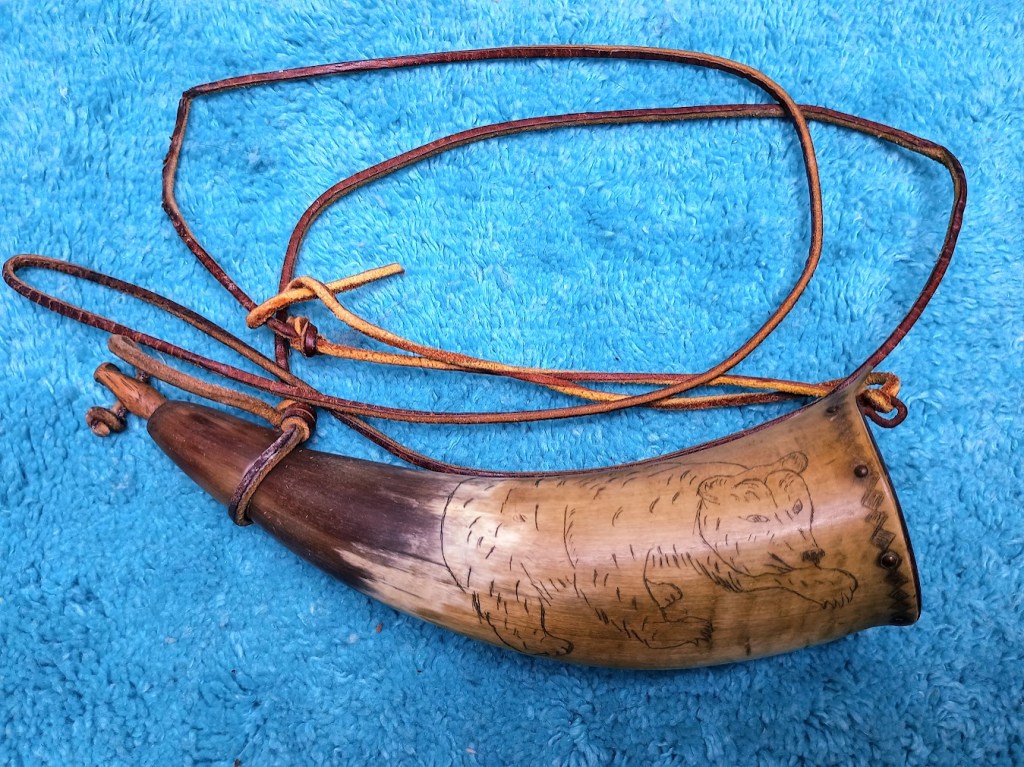An intricately carved powder horn featuring a depiction of a bear, secured with leather straps, placed on a blue textured background.