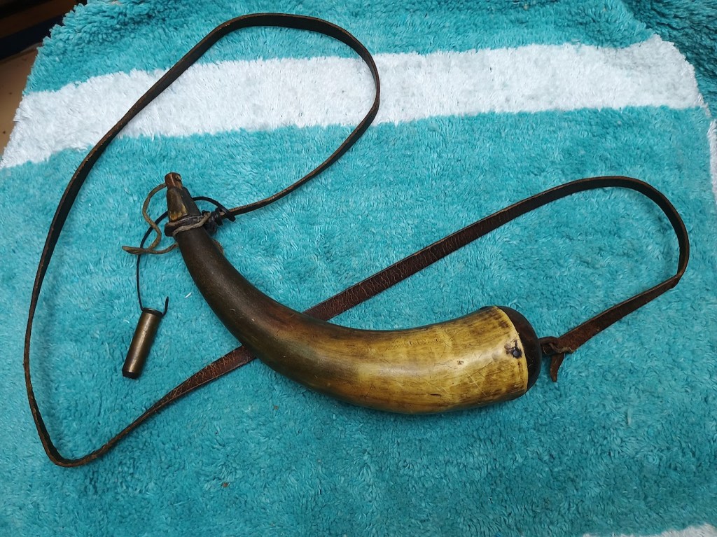 A traditional powder horn made from a hollowed-out bull horn with a leather strap and a wooden stopper, resting on a blue towel.